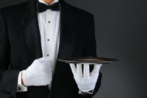 Closeup of a tuxedo wearing waiter holding a silver tray in front of his body. Horizontal format on a light to dark gray background.